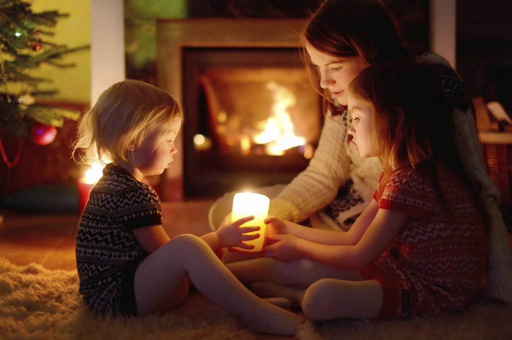 Young mother and her two little daughters sitting by a fireplace holding candles in a cozy dark living room on Christmas eve