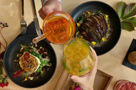 Man sieht einen gedeckten Tisch von oben, über dem die Hände von zwei Personen mit Getränken anstoßen.