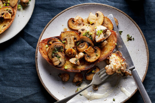 Zu sehen ist ein mit Pilzen belegtes, gebackenes Brot auf einem Teller mit Besteck.