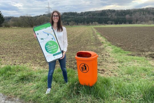 Michaela Heigl auf einem Feld vor einem Mülleimer, eine große Werbung für die App Digi-Cycle in Händen.