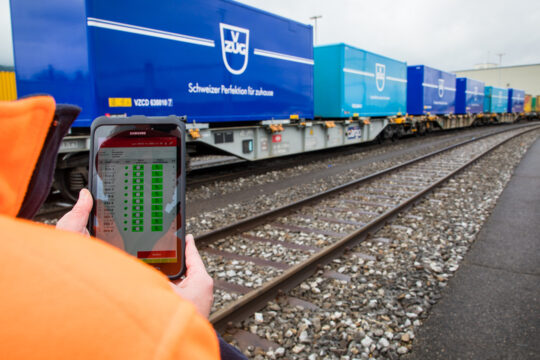 Eine Person hält eine digitales Gerät vor Zugschienen und Güterzügen.