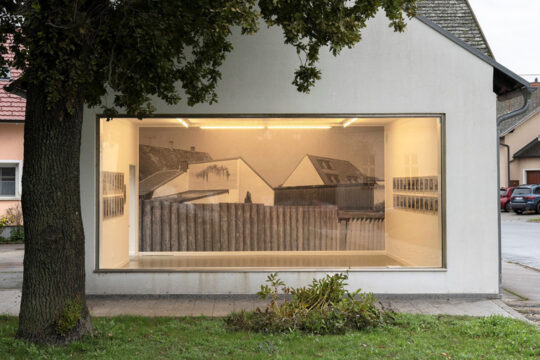 Ein kleines Haus mit einer Glaswand, durch die man ins Innere sehen kann. Dort ist eine Ausstellung.