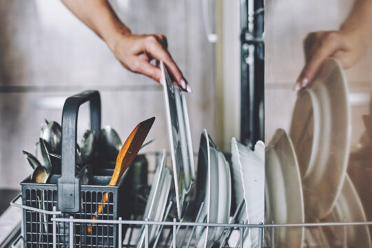 Eine offene Geschirrspülmaschine von der Seite, in die eine Hand einen Teller einschlichtet.