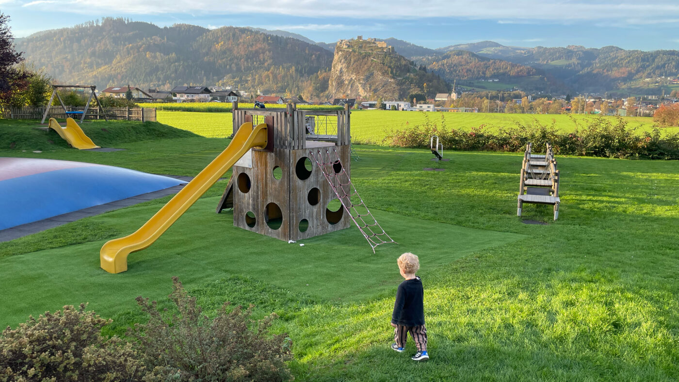 Ein Kind auf einer grünen Wiese vor einem Spielplatz.