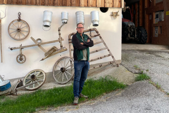 Eine Person steht mit verschränkten Armen vor einem Hof, daneben führt ein offenes Tor in das Gebäude.