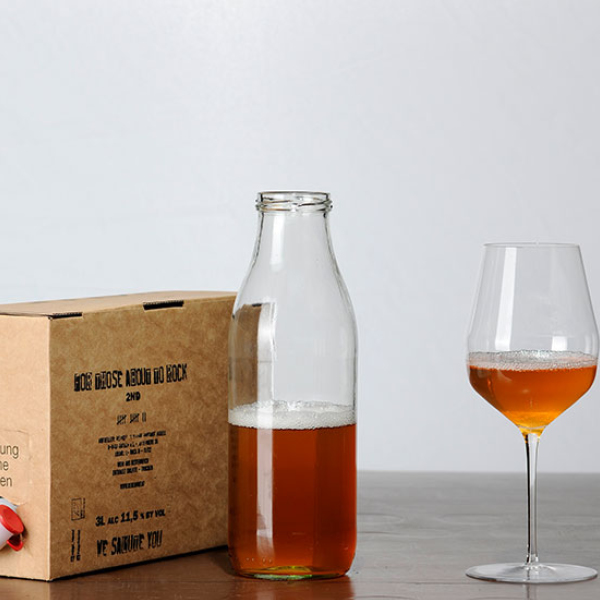 Neben einem Karton stehen eine Glasflasche und ein Weinglas mit einer rötlichen Flüssigkeit gefüllt.