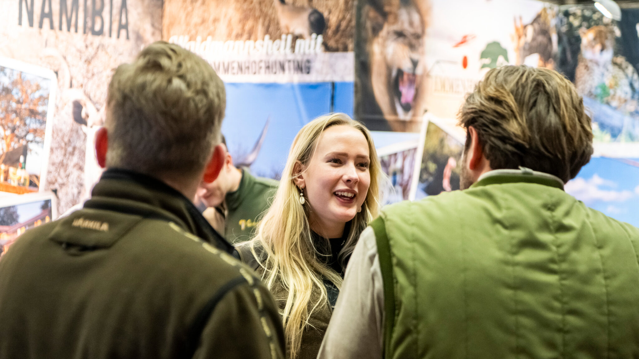 Mehrere Personen stehen auf einer Jagdmesse. Auf Bildern im Hintergrund sind Angebote für Jagd auf Löwen zu sehen.