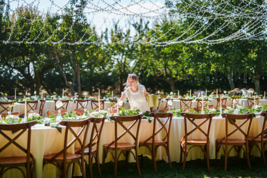 Eine Person im weißen Kleid steht vor einem aufwändig gedeckten Tisch für viele Menschen im Freien.