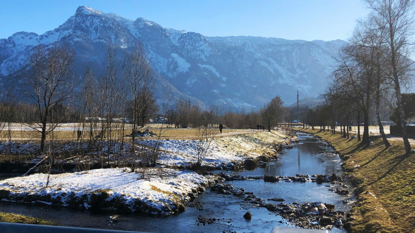 Ein Fluss mit schnee an der Böschung vor einem Bergpanorama.