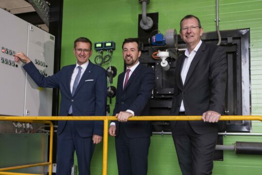 Drei Menschen in Anzügen stehen in einem Raum, neben ihnen eine Maschine auf der eine Person einen Knopf drückt.