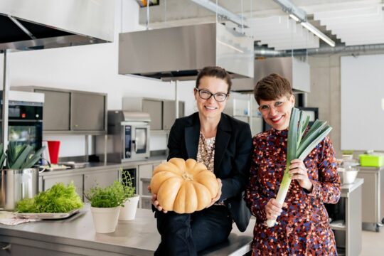 Zwei Personen in einer großen Küche, eine hält einen großen Kürbis, die andere einen Lauch.