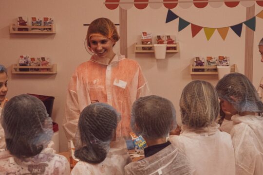 Im Bild zu sehen ist eine unbekannte Person bei einer Führung vor 5 Kindern, deren Köpfe von hinten zu sehen sind.