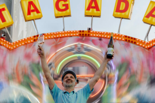 Ein Mann steht vor einem Tagada (Kirmes-Fahrgeschäft) und hält zwei Flaschen hoch.