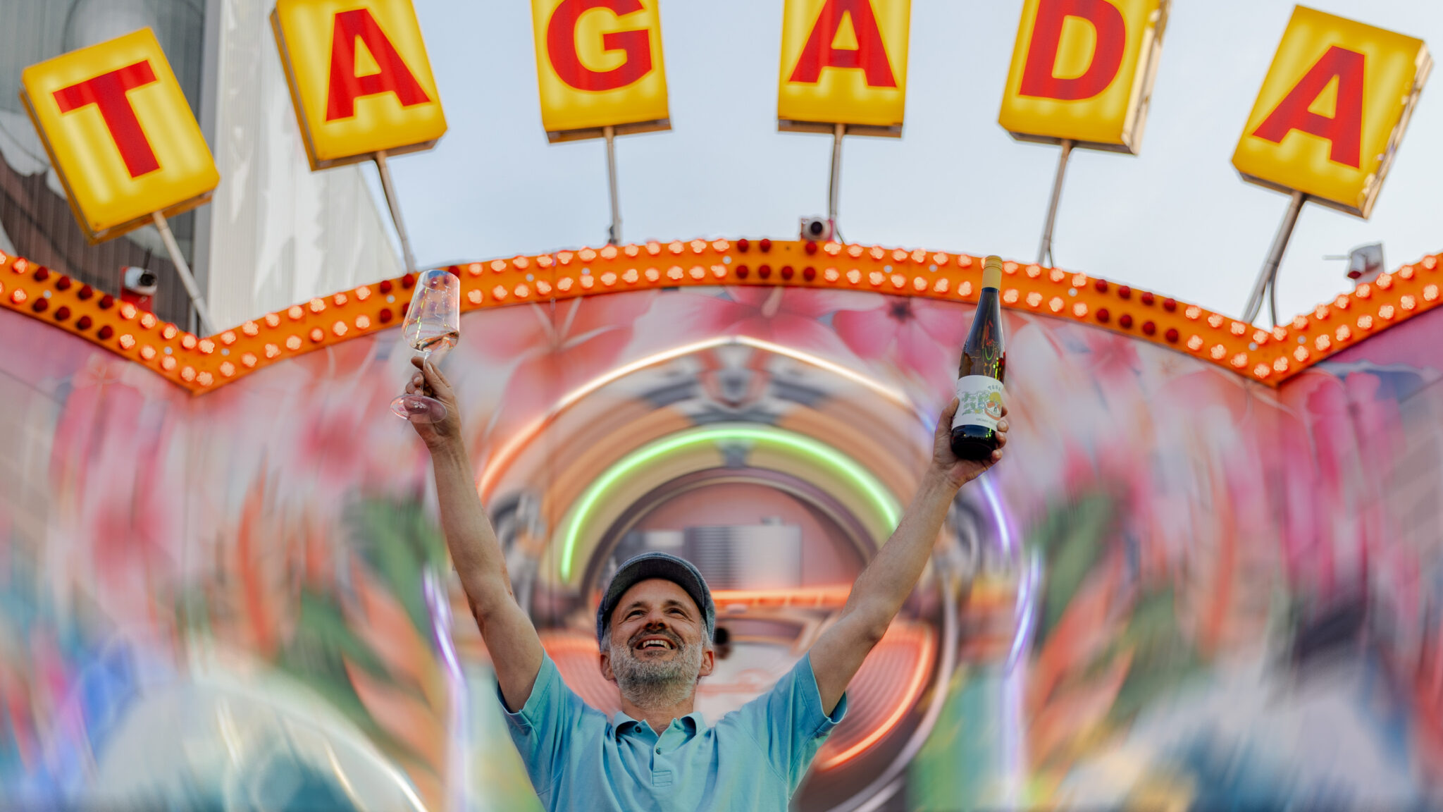 Ein Mann steht vor einem Tagada (Kirmes-Fahrgeschäft) und hält zwei Flaschen hoch.